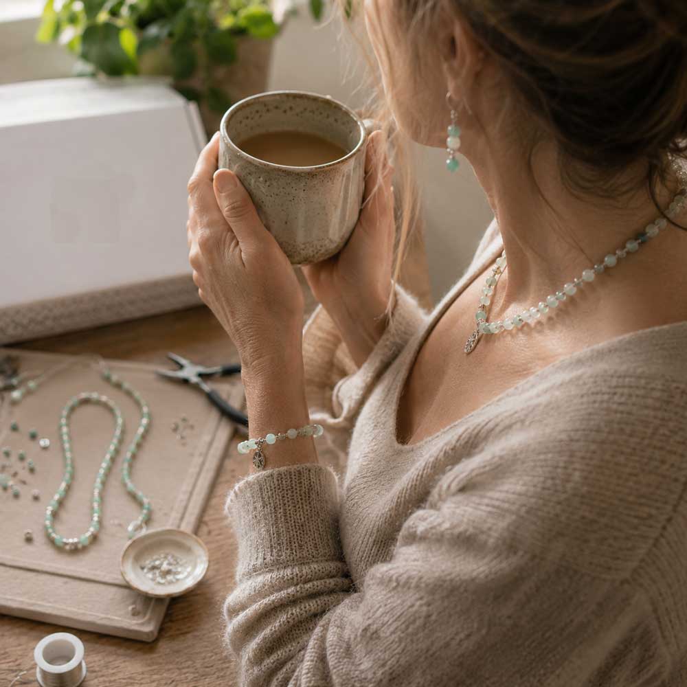 Finished handmade beaded necklace on a rustic craft table beside jewellery-making tools, while a maker relaxes with a cup of tea after completing the project.